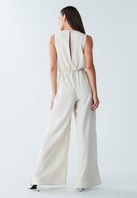 Beige jumpsuit med en struktureret halsudskæring, åben rygdetalje, elastik i taljen og vidde ben. Glat stof med subtil struktur.