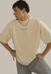 Beeži värvi lahtine t-särk, millele on tumeda niidiga tikitud sõnad „Luxury Chaos“, kantud koos helesiniste teksapükstega; minimaalsete aksessuaaridega.