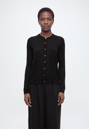 Rövid hajú nő, fekete, testhezálló, gombos kardigánban és laza fekete nadrágban, sima világos háttér előtt áll.