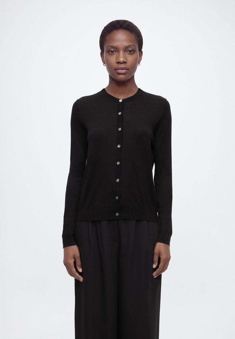 Rövid hajú nő, fekete, testhezálló, gombos kardigánban és laza fekete nadrágban, sima világos háttér előtt áll.