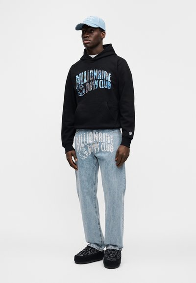 Μαύρο φούτερ με κουκούλα και μπλε γραφικό κείμενο, ανοιχτόχρωμο τζιν παντελόνι με τυπωμένο λογότυπο που ταιριάζει, μαύρα κεντημένα παπούτσια χωρίς κορδόνια, ανοιχτόχρωμο καπέλο.