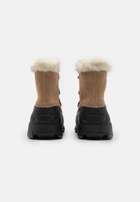 Brune suedeboots med sort gummisåle og faux pelskrage, rund tå, snøring foran og mønstret såle, med Sorel-merke på siden.