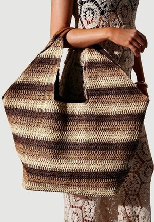 Braccio che tiene una grande borsa a uncinetto a righe nei toni del marrone e beige, indossando un vestito a uncinetto beige e un braccialetto dorato, sfondo neutro.