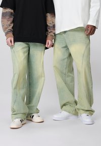 Δύο άτομα που φορούν φανταχτερά χακί φαρδιά τζιν, το ένα συνδυασμένο με λευκά αθλητικά παπούτσια με μαύρες ρίγες, το άλλο με απλά λευκά αθλητικά παπούτσια.