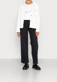 Chaqueta de mezclilla blanca sobre una camiseta gráfica blanca, combinada con jeans de pierna ancha de talle alto negros y zapatos negros robustos sobre un fondo neutro.