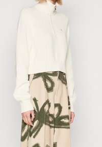 Kvinna klädd i en krämfärgad stickad tröja med halvdragkedja och beige kordfloreet byxor med stora abstrakta mörkgröna mönster.