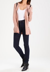 Light pink cardigan with open front, textured fabric, and black shoulder accents. Paired with dark skinny jeans and pointed black ankle boots.