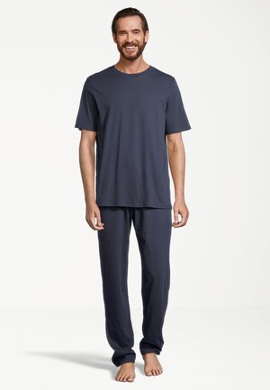 Uomo in piedi a piedi nudi che indossa una camicia blu scuro a maniche corte abbinata e pantaloni lunghi, sorridente su uno sfondo chiaro e uniforme.