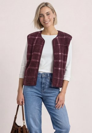 Frau trägt eine burgunderrote karierte Fleeceweste über einem weißen Langarmshirt und blauen Jeans und hält eine braune Lederhandtasche.