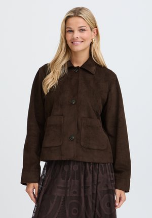 Femme souriante aux longs cheveux blonds, portant une veste boutonnée marron foncé avec de grandes poches devant et une jupe marron à motif.