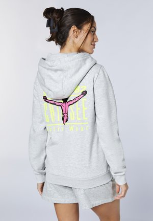Young woman with dark hair in clip wearing light gray hoodie and shorts, facing away showing colorful graphic on hoodie back.
