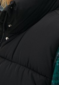 Černá prošívaná vesta s vysokým límcem, vybavená patentními knoflíky a prošívanou strukturou. Materiál je hladký a izolovaný, ideální pro teplo.