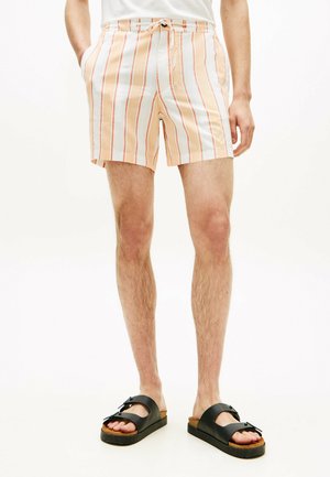 Hombre con pantalones cortos a rayas blancas y naranjas y sandalias negras abiertas en los dedos, de pie con las manos en los bolsillos sobre un fondo blanco.