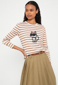 Pruhovaná tričko s dlhým rukávom v béžovej a bielej farbe, s kresleným obrázkom mačky. Tričko má okrúhly výstrih a z jemného bavlneného materiálu.