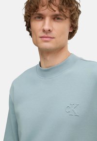 Sudadera de color azul claro, confeccionada en una mezcla de algodón, con un cuello redondo acanalado y un logo en relieve en el lado izquierdo del pecho. Textura suave.