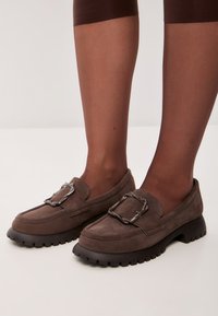 Mocassini in suede marrone con suola in gomma nera chunky, caratterizzati da un accento di catena metallica sulla parte superiore. Superficie testurizzata con cuciture a contrasto.