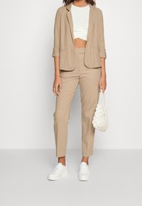 Beżowa lniana marynarka na białym krótki top, w zestawie z beżowymi spodniami i białymi trampkami. Torba jest biała i ma fakturę.