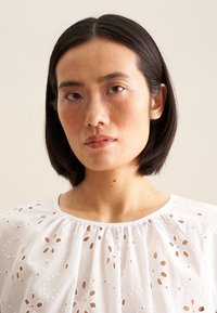 Blusa bianca con intricate fantasie a occhio di pernice, scollatura rotonda e tessuto morbido e fluido. Carnagione naturale con trucco minimal.
