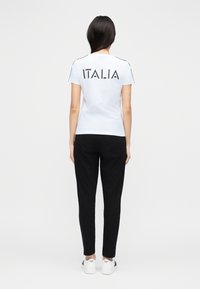 Balta īsroku T-krekls ar uzrakstu "ITALIA" uz muguras, kombinēts ar melnām plānām biksēm un baltām sporta kurpēm.