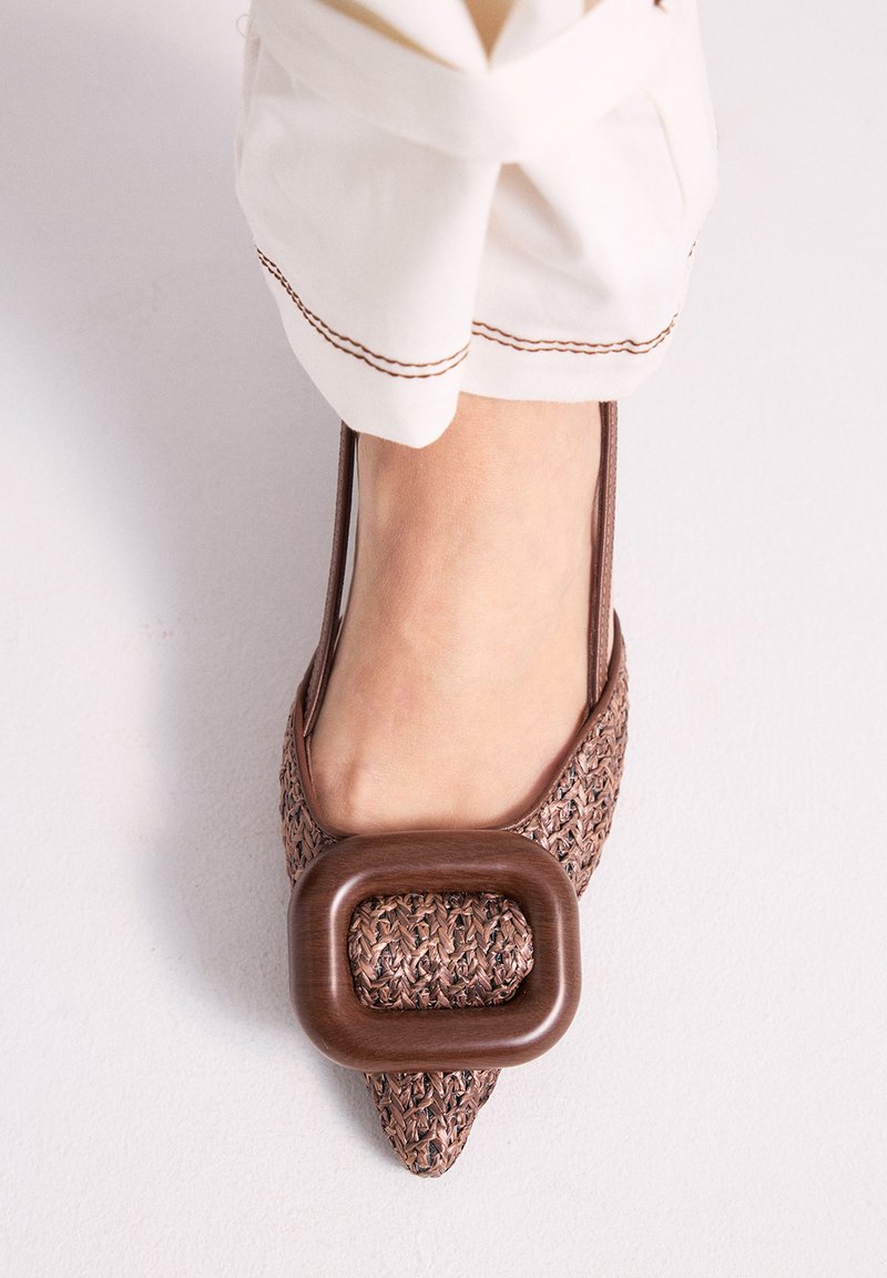 Pie con zapato plano puntiagudo marrón tejido con una gran hebilla rectangular, combinado con pantalones blancos que presentan costuras marrones en los puños.