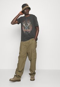 Camiseta gráfica negra con diseño de serpiente, combinada con pantalones cargo olivo y zapatos beige. Accesorio: sombrero tipo bucket en beige y marrón.