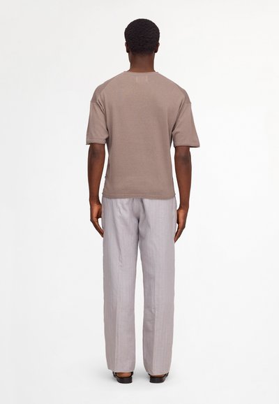 Homme debout, de dos, portant une chemise marron à manches courtes, un pantalon ample gris clair et des chaussures noires sur un fond uni.