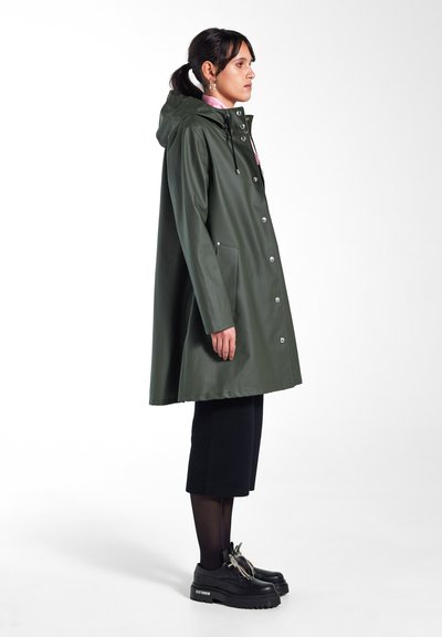 Manteau de pluie long, vert olive, en matériau imperméable, avec une capuche, des pressions argentées et des poches latérales. Associé à des chaussures à plateforme noires et un pantalon.