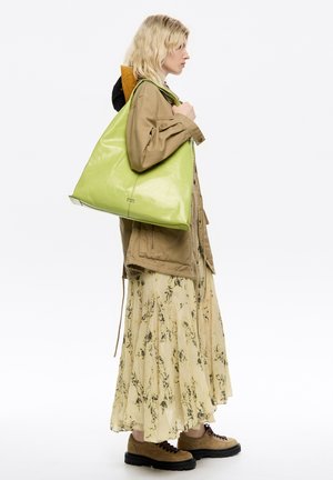 Zielona skórzana torba o trójkątnym kształcie, błyszczącej teksturze i detalach logo u podstawy, noszona na ramieniu przez osobę w casualowym stroju.