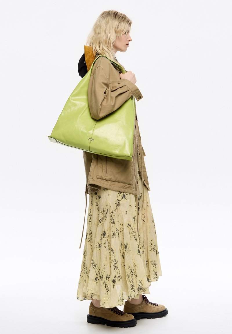 Zielona skórzana torba o trójkątnym kształcie, błyszczącej teksturze i detalach logo u podstawy, noszona na ramieniu przez osobę w casualowym stroju.