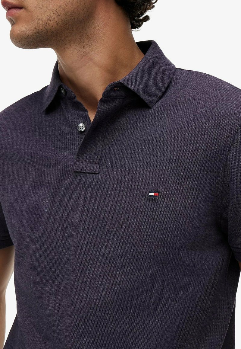 Homme portant un polo violet foncé avec deux boutons et un petit logo rectangulaire rouge, blanc et bleu sur la poitrine gauche.