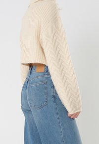 Pull tricoté couleur crème avec un design court et un motif en zigzag texturé, associé à un jean en denim bleu taille haute.