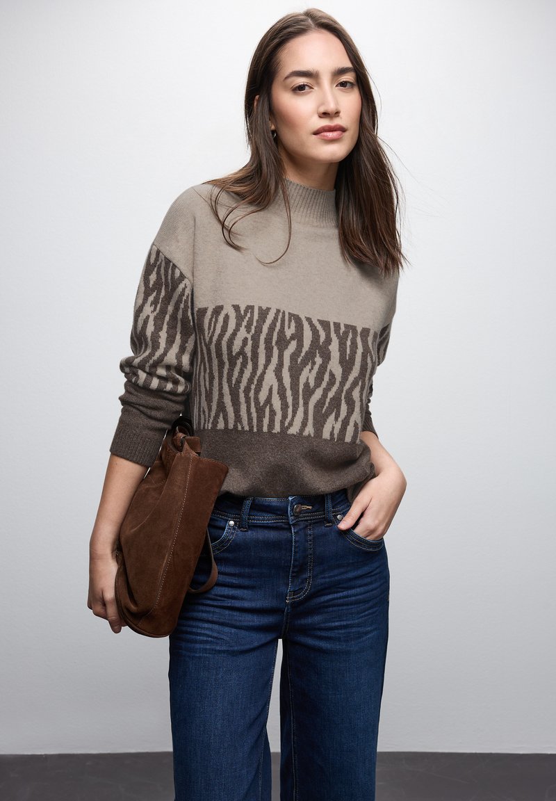Beiger und brauner Strickpullover mit einem Zebra-Streifenmuster über der Brust. Kombiniert mit blauen Jeans und einer braunen Wildlederhandtasche.