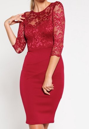 Woman wearing a fitted knee-length red dress with lace three-quarter sleeves and sheer lace yoke detailing.