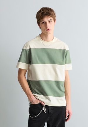 Jeune personne aux cheveux bruns courts portant un t-shirt rayé vert et crème et un pantalon noir avec des clés accrochées à une chaîne.