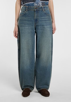 Osoba nosząca luźne, wyblakłe niebieskie dżinsy z przednimi kieszeniami i zapięciem na guziki, w zestawieniu z brązowymi wsuwanymi butami na tle jednolitego tła.