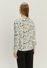 Długi rękaw bluzki w kremowym materiale z czarnym wzorem kwiatowym w zarysie. Design jest luźny, z miękką teksturą i okrągłym dekoltem.