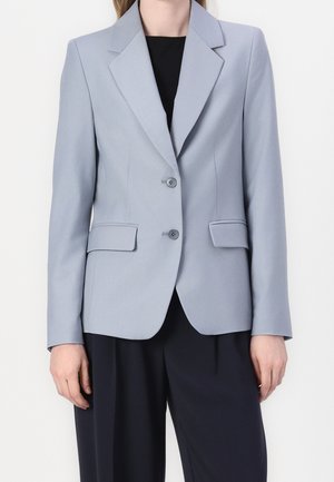 Femme portant un blazer gris clair ajusté avec deux boutons, poches à rabat, et pantalon noir sur fond blanc.