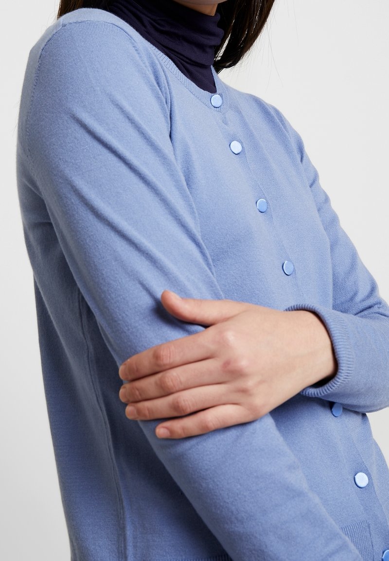 Světlounce modrý svetr na zapínání z měkké tkaniny, s kulatým výstřihem, dlouhými rukávy a modrými knoflíky vpředu. Hladká textura.