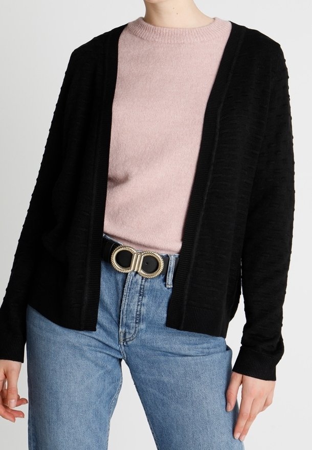 Femme portant un pull rose clair, un cardigan noir ouvert, un jean bleu taille haute et une ceinture noire avec une boucle double anneau dorée.