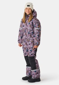 Paarse skipak met dierenpatronen, zwarte accenten, reflecterende strepen en bijpassende broek. Draagt een witte gebreide muts en zwarte laarzen.