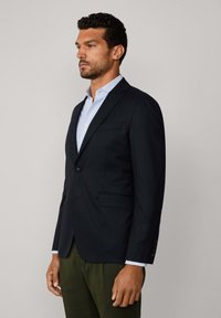 Veste en laine bleu marine avec un bouton unique, revers crantés, deux poches avant et une coupe élégante. Portée sur une chemise bleu clair.