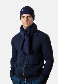 Mann trägt eine marineblaue Strickmütze, dazu passend ein im Vordergrund gebundener Schal, eine gefütterte Jacke und hat die Hände in den Taschen vor einem schlichten Hintergrund.
