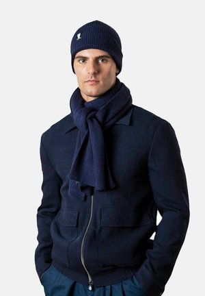 Mann trägt eine marineblaue Strickmütze, dazu passend ein im Vordergrund gebundener Schal, eine gefütterte Jacke und hat die Hände in den Taschen vor einem schlichten Hintergrund.