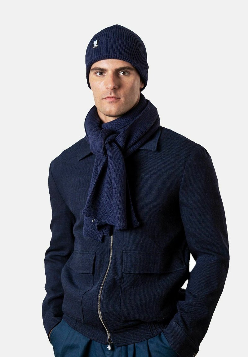 Mann trägt eine marineblaue Strickmütze, dazu passend ein im Vordergrund gebundener Schal, eine gefütterte Jacke und hat die Hände in den Taschen vor einem schlichten Hintergrund.