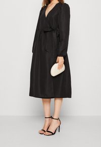 Kvinna i en svart omlottklänning med långa ärmar, som håller en vit clutch, bär svarta högklackade sandaler och lager-på-lager halsband.