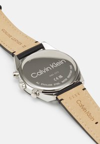 Montre en acier inoxydable avec un dos argenté poli, un bracelet en cuir noir et un logo de marque gravé ; étanche jusqu'à 3 bars.