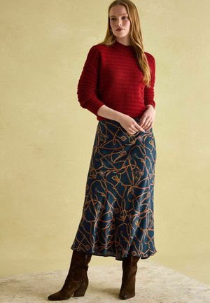 Roter Strickpullover mit langen Ärmeln, kombiniert mit einem langen marineblauen Rock mit braunem geometrischem Muster, ergänzt durch braune kniehohe Stiefel.