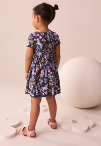 Klänning med blommönster i marinblått med korta ärmar och volangkant, kombinerad med rosa sandaler. Slät textur, avslappnad design.