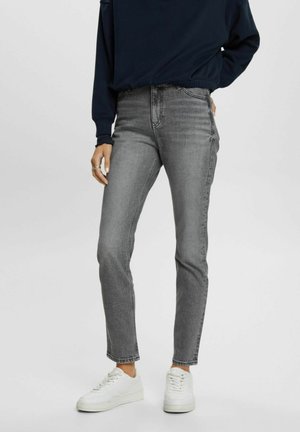 Personne portant un jean slim gris, des baskets blanches et un haut à manches longues foncé, debout devant un fond uni.