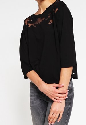 Kobieta ubrana w czarny top z przezroczystymi kwiatowymi detalami na górnej części klatki piersiowej oraz rękawami do połowy przedramienia, zestawiony z szarymi jeansami denimowymi.